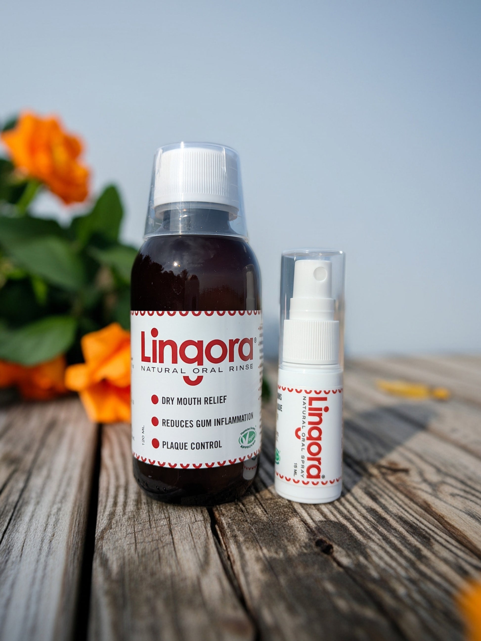 A bottle of Lingora natural oral rinse and a matching spray bottle placed on a weathered wooden surface with blurred orange flowers and a pale blue sky in the background.